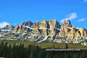 A brown mountain with the peaks cut off, an evergreen forest and snow in front of the moutains, and a dark blue sky with a few thin fluffy white clouds