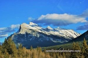 A large jagged mountain with an evergreen forest in front of it and blue sky with large fluffy white clouds