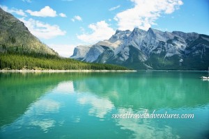 Blue cloudy sky, grey mountains, and green grass reflecting on calm blue lake