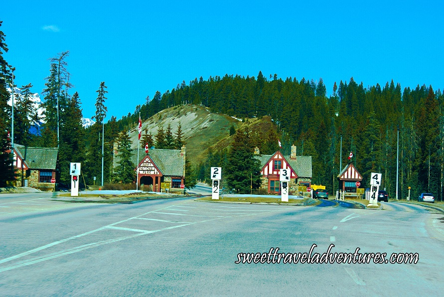 Road with four lanes and a small brown wooden building next to each lane, each building has a Canadian flag on the top of it, evergreen trees and mountains in the background, and a clear blue sky except for one small and faint white cloud on the left-hand side