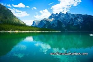 Blue cloudy sky, grey mountains, and green grass reflecting on a calm blue lake