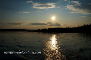 Low-lying sun in blue and light orange sky with several large fluffy white clouds all reflected on rippled water with shadowed trees in the background