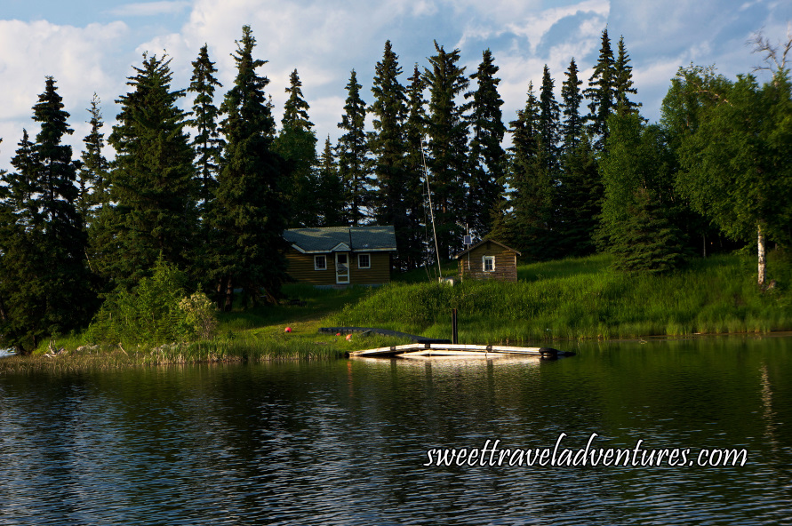 A wooden cabin nestled between trees with a miniature cabin to the right on a grassy shore with a small dock, rippled water, and a blue sky filled with large fluffy white clouds