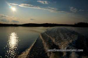 Sun starting to set in blue and light orange sky with several large fluffy white clouds all reflected on water which has a wake from a boat and trees shadowed in the background