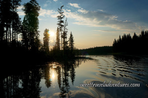 Sun starting to set in blue and light orange sky with a few fluffy white clouds and shadowed trees all reflected on the lake which also has a wake pattern from the boat