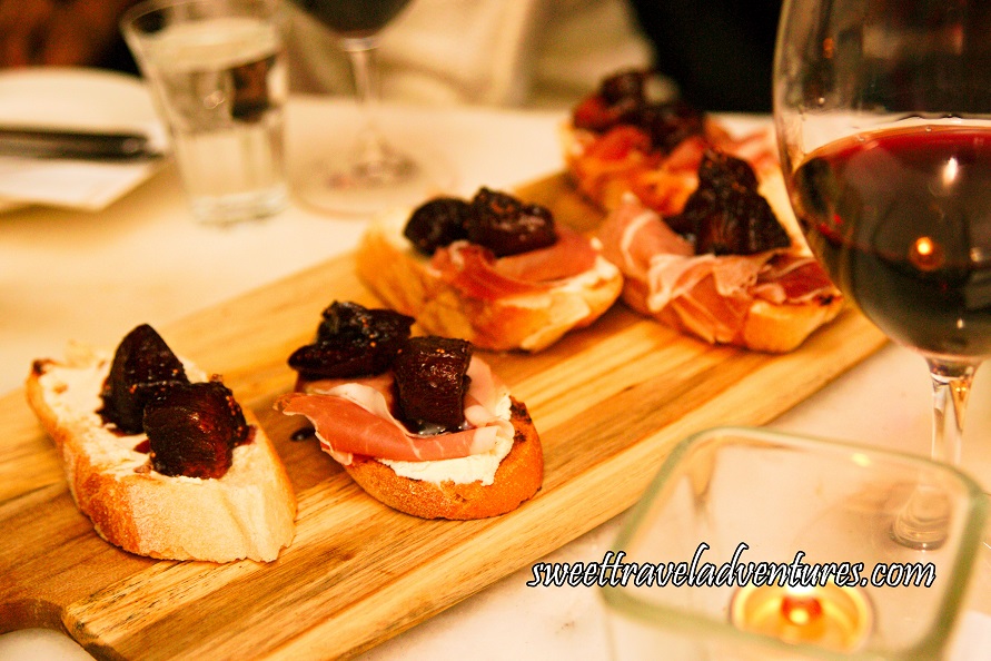 A Wooden Cutting Board Angled on a Table With Five Slices of Grilled Baguette, Each With Two Pieces of Fig on Top of Prosciutto, Which is on Top of Mascarpone and a Tealight Candle in a Square Glass Votive as well as a Glass of Red Wine Next to the Wooden Board