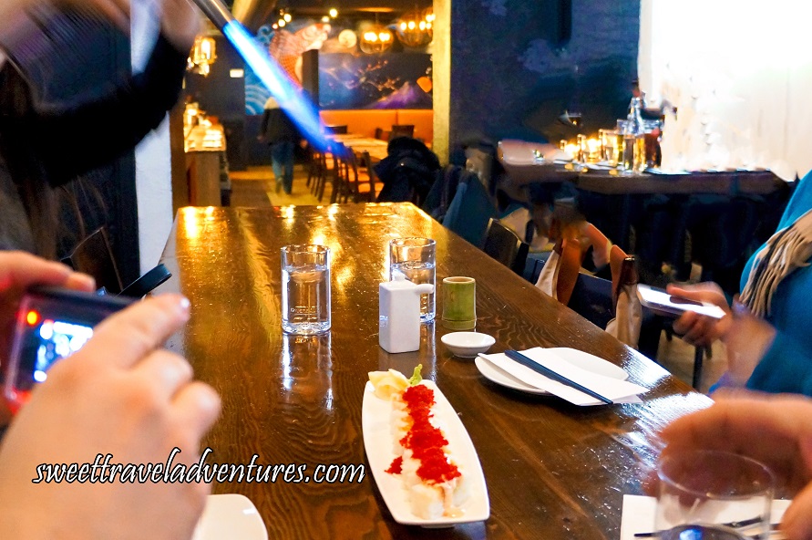 9 or 10 Pieces of Sushi With Red Flying Fish Roe on Top of Each Piece and Placed on a Long White Rectangular Dish, on a Long Rectangular Wooden Table, Being Torched 