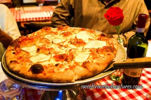 A Pizza Server Placed Under a Pizza With Thin Slices of Pear and Covered in Cheese Sitting on a Silver Platter Stand on a Table With a Red and White Checkered Table Cloth Next to a Bottle of Wine and a Red Rose Placed in an Old Glass Coke Bottle