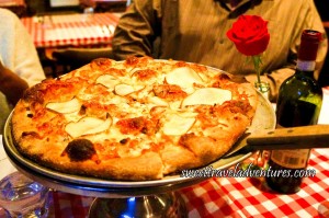 A Pizza Server Placed Under a Pizza With Thin Slices of Pear and Covered in Cheese Sitting on a Silver Platter Stand on a Table With a Red and White Checkered Table Cloth Next to a Bottle of Wine and a Red Rose Placed in an Old Glass Coke Bottle
