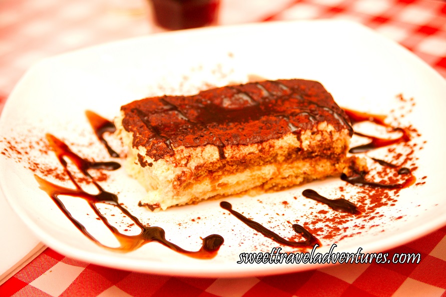 A Fluffy White Rectangular Dessert With a Dark Brown Layer on Top, on A White Plate With Golden Brown Liquid Swirls Around the Plate and Some Golden Brown Liquid Across the Top of the Dessert