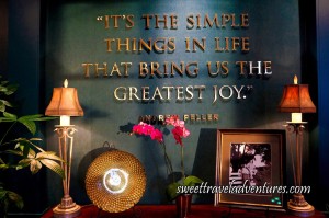 A Quote in Gold Lettering Saying It's the Simple Things in Life That Brings Us the Greatest Joy on a Dark Green Wall With Decorations on a Wooden Table Below the Quote