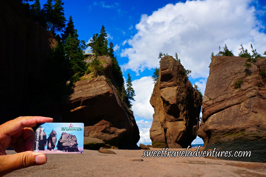 A Blue Sky With Two Huge Fluffy White Clouds and Three Huge Brown Rocks With the Middle One Facing Us Which Looks Like an Elephant Except the Trunk is Missing and There is a Sandy Brown Floor, the View is the Same as the Picture on a New Brunswick Health Care Card that is Being Held Next to it
