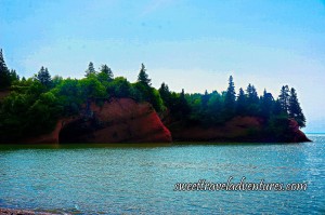 Large Reddish Brown Rock With an Opening and Green Trees Growing Out of the Top of the Rock, Fairly Calm Blue Water and a Light Blue Sky