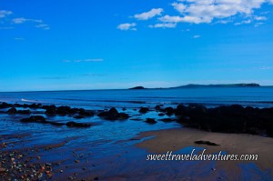 Blue Sky With a Few Fluffy White Clouds and Blue Ocean Water, Land in the Distance Covered in Greenery, A Few Small Waves Close to Shore, Water Washing Up on the Sandy Beach, Large Rocks Covered in Black Seaweed on the Beach With A Big Cluster of Them to the Right, Some Pebbles of Different Colours to the Left
