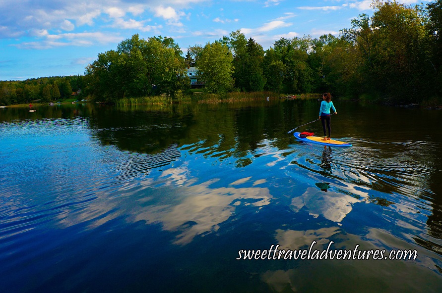 Blue Sky With Small Fluffy White Clouds Reflected Onto the Lake, Someone Standing on a Long Blue and Yellow Board With Their Back Towards Us Holding a Long Paddle, Reeds, Green Grass, and Green Trees Around the Lake and a House Peeking Out Between the Trees in the Middle