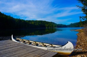 A Bright Blue Sky With Streaks of White, Several Green Trees Around a Blue Lake and Reflecting on it, a Wooden Dock Angled to the Left With a Long White Canoe Next to it, the Edge of a Shore to the Right With Dirt and Rocks