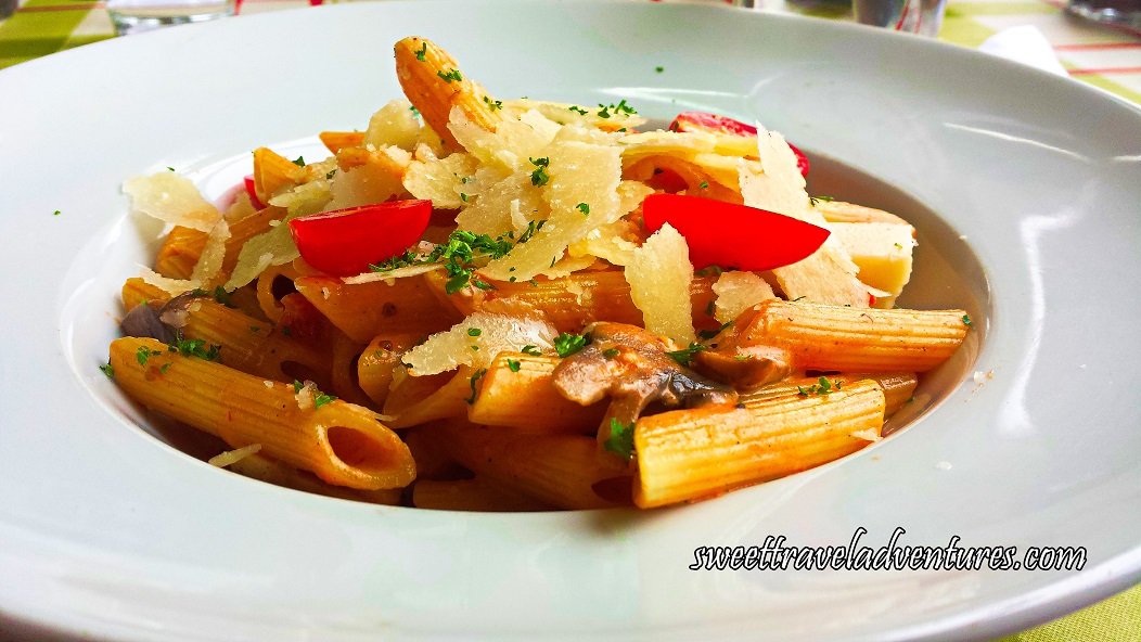 Penne With Mushrooms, Half-Cut Cherry Tomatoes, Pieces of Parmesan Cheese, and Herbs in a White Pasta Bowl on a Table With a Checkered White, Red, and Green Tablecloth