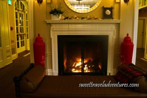 A Large Fireplace With Glass Doors, White Molding Around it and a White Mantle, a Round Mirror Above the Fireplace on an Off-White Wall, a Brown Leather Lounge Chair in Front of the Fireplace With a Red Checkered Throw