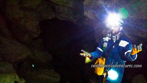 A Cave Opening of Brown Rock With a Man Standing on the Right of the Opening in Dark Blue Coveralls and a Helmet With a Headlight Turned On