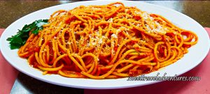 A Spaghetti-Like Pasta But Thicker With a Red Sauce, Sprinkled With White Cheese, and a Garnish of Parsley Leaves on the Left, on Top of a White Plate That is Sitting on a Brown Table