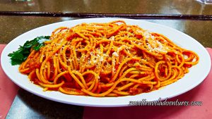 A Spaghetti-Like Pasta But Thicker With a Red Sauce, Sprinkled With White Cheese, and a Garnish of Parsley Leaves on the Left, on Top of a White Plate That is Sitting on a Brown Table