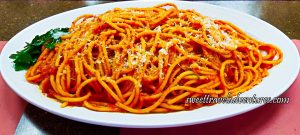 A Spaghetti-Like Pasta But Thicker With a Red Sauce, Sprinkled With White Cheese, and a Garnish of Parsley Leaves on the Left, on Top of a White Plate That is Sitting on a Brown Table