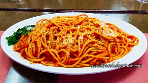 A Spaghetti-Like Pasta But Thicker With a Red Sauce, Sprinkled With White Cheese, and a Garnish of Parsley Leaves on the Left, on Top of a White Plate That is Sitting on a Brown Table