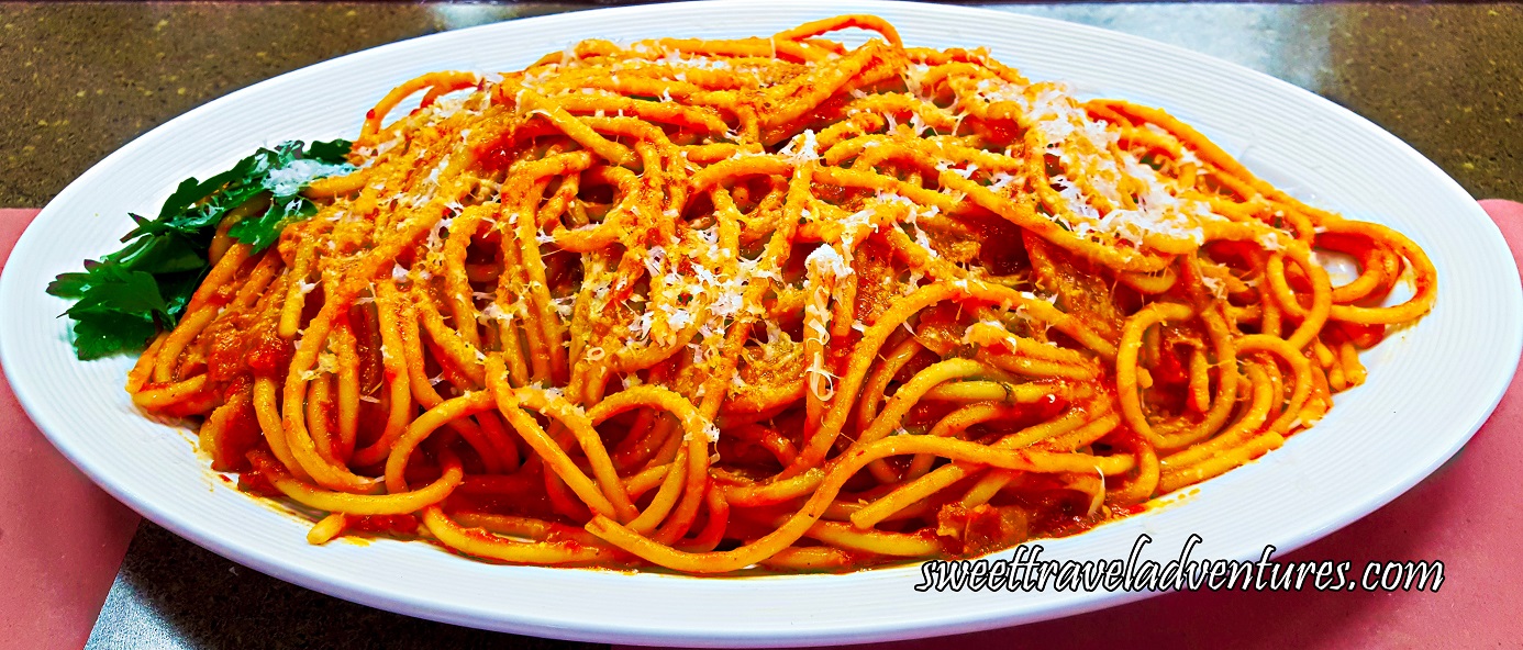 A Spaghetti-Like Pasta But Thicker With a Red Sauce, Sprinkled With White Cheese, and a Garnish of Parsley Leaves on the Left, on Top of a White Plate That is Sitting on a Brown Table