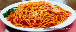 A Spaghetti-Like Pasta But Thicker With a Red Sauce, Sprinkled With White Cheese, and a Garnish of Parsley Leaves on the Left, on Top of a White Plate That is Sitting on a Brown Table