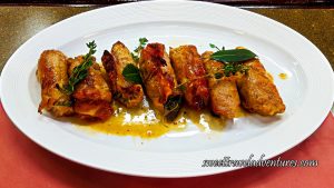 Pork Rolled With Prosciutto on the Outside and Stuffed Inside, With a Little Sauce on a White Plate on a Table and Garnished With Herbs