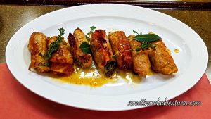 Pork Rolled With Prosciutto on the Outside and Stuffed Inside, With a Little Sauce on a White Plate on a Table and Garnished With Herbs