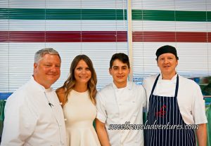 A Man, A Woman, and Two Men All Standing in Front of White Blinds With a Red, White, and Green Stripe at the Top (Like the Italian Flag), the Men are Wearing White Chef Jackets and the Man on the Far Right is Wearing a Black and White Vertically Striped Apron on Top of His Chef Jacket, and the Woman is Wearing a White Sleeveless Blouse