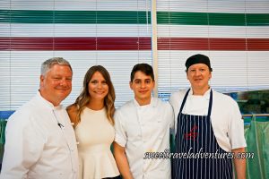 A Man, A Woman, and Two Men All Standing in Front of White Blinds With a Red, White, and Green Stripe at the Top (Like the Italian Flag), the Men are Wearing White Chef Jackets and the Man on the Far Right is Wearing a Black and White Vertically Striped Apron on Top of His Chef Jacket, and the Woman is Wearing a White Sleeveless Blouse