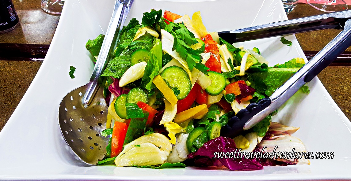 Salad of Romaine Lettuce, Red and Yellow Peppers, Red Cabbage, Cucumber, Tomatoes, and Parsley in a White Dish With a Large Silver Spoon and Silver Tongs and the Dish is on a Brown Table