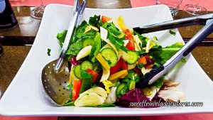 Salad of Romaine Lettuce, Red and Yellow Peppers, Red Cabbage, Cucumber, Tomatoes, and Parsley in a White Dish With a Large Silver Spoon and Silver Tongs and the Dish is on a Brown Table