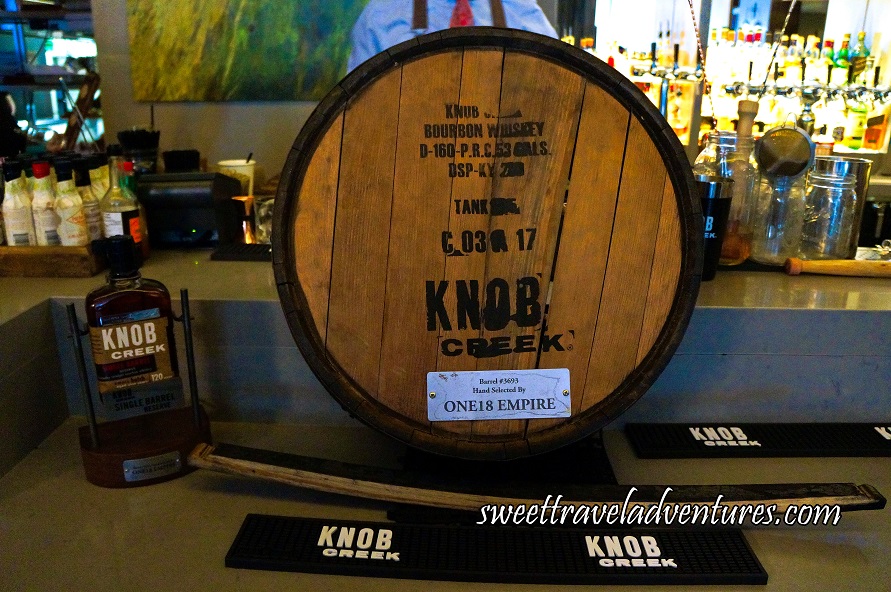 The Bottom of a Wooden Barrel Sitting on a Grey Bar Stamped in Black Ink and a Metal Label, in Front is a Piece of the Side of the Barrel, on the Left is a Bottle on a Stand, and Bottles and Mixology Equipment Behind