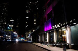 One18 Empire on the Right Next to a Sidewalk and Street (A Black Building With White Lettering and White Columns and a Few Rectangles Lit up in Raspberry and Purple), Short White Buildings Next to it, and Tall Office Buildings With a Few Lights on in the Background
