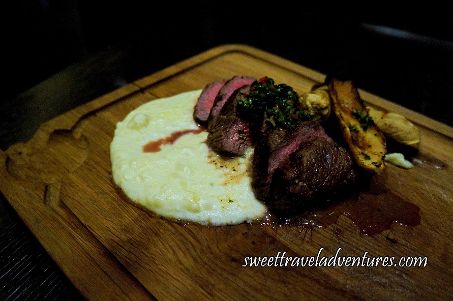 A Medium-Coloured Wooden Cutting Board Sitting on an Angle on a Dark Brown Wooden Table With Slices of Medium Cooked Meat Topped With a Green Chimichurri, to the Right is Sliced Grilled Mushrooms, Underneath and to the Right is Mashed Potatoes
