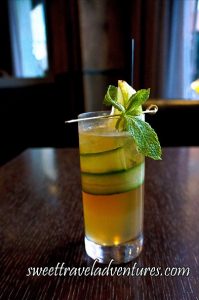 A Drink in a Tall Glass on a Dark Wooden Table With a Golden Yellow Liquid and Strip of Cucumber Around the Top Inside of the Glass, A Metal Pick Sitting on the Top of the Glass With a Mint Leaf, and a Lemon Wedge Sticking Out of the Top of the Glass Along With a Long Black Straw Behind it