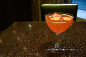 A Drink in a Cocktail Glass Sitting on a Dark Wooden Table With a Dark Brown Leather Chair Behind and a Portion of a Window on the Right With the Road and Sidewalk, the Glass is Filled With a Peachy Pink Liquid and Floating on Top is Three Slices of Strawberry