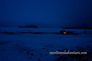 A Couple at a Campfire at Dusk With Snow Around Them and Trees in the Background and a Dark Blue Sky