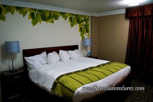 A Bed With a Cherry Wood Frame and Night Tables Next to it With White Lamps, the Sheets on the Bed are White and a Bright Green Blanket on the Lower Portion of the Bed, a White Wall With Rich Green and Bright Green Leaves Bordering it, on the Right is a Tan Coloured Wall With Reddish Brown Satin Curtains, and a Patterned Carpet