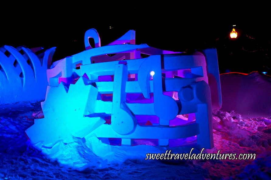 A Snow Sculpture of Musical Notes and a Large Maple Leaf on the Left, All Lit Up at Night With Blue and Purple Lights