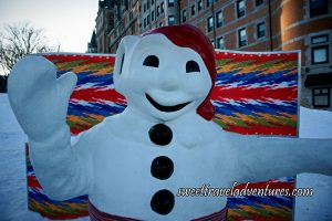 The Upper Half of a Snowman Mascot Wearing a Red Touque and a Sash Around its Waist, a Multi-Coloured Fabric Behind, and a Hotel in the Background