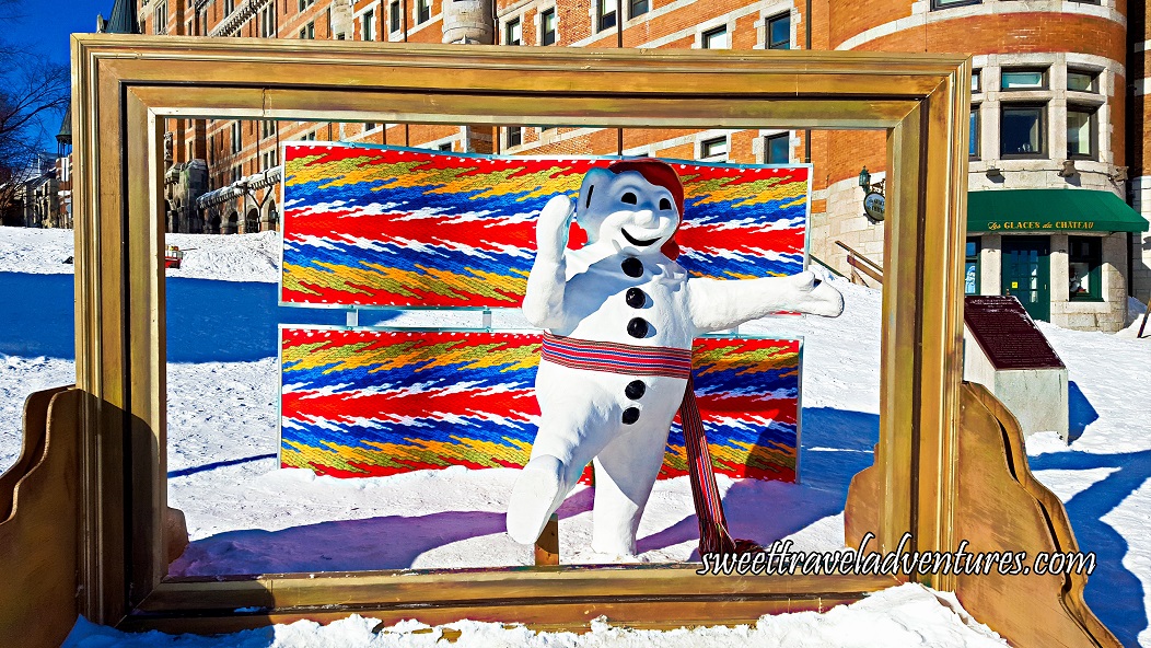 A Wooden Picture Frame With a Snowman Mascot Standing and Waving Inside the Frame Wearing a Red Tourque and a Colourful Sash, Directly Behind is Two Large Pieces of Material Like the Sash, in the Background is a Large Hotel, and the Ground is Covered in Snow