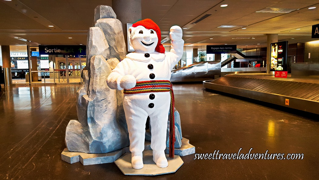 At an Airport is A Mascot Statue of a Snowman Wearing a Red Touque and Multi-Coloured Sash Around its Waist and Giant Rocks Behind it, on the Right is a Baggage Carousel, and Behind in the Distance is Glass Exit Doors