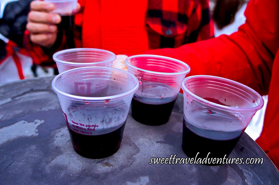 Plastic Cups Filled Almost Halfway With a Dark Red Liquid Sitting on a Grey Tray With a Person in a Red Jacket Holding the Tray on the Right and a Person Behind Holding One of the Cups of Dark Red Liquid Wearing a Red and Black Checkered Shirt and Red Scarf