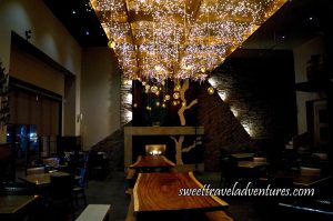 A Room With Two Long Wooden Tables in the Middle With Many Tiny Lights Hanging Above and Some Hanging Balls With Lights Inside, a Fireplace With Stone Lit in Front of Them, on Either Side Dark Wooden Tables With Matching Wooden Chairs With Metal Embellishments and Glass Windows on the Left