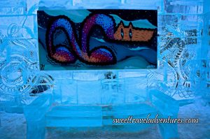 An Ice Wall With Intricate Designs With a Painting Hanging on it and a Bench Made of Ice Below and the Ground Lightly Covered in Snow