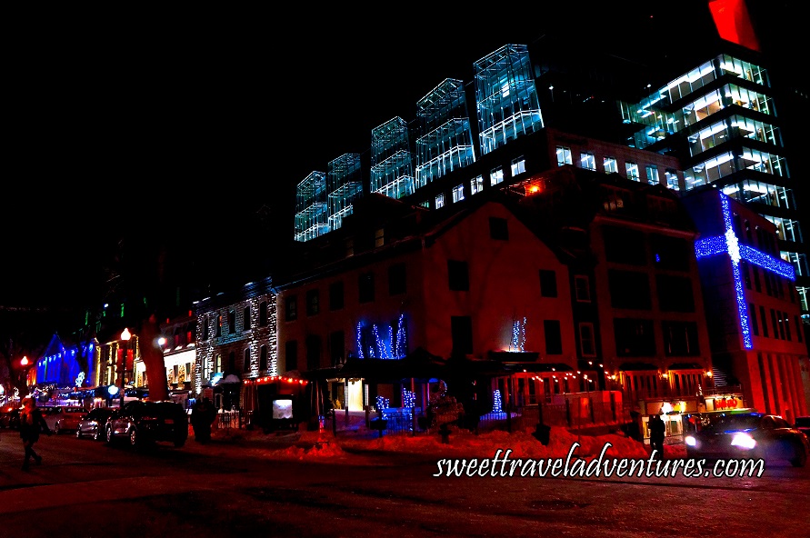 A Street at Night With the Buildings Lit Up With Dark Blue, Green, White, or Red Lights, and a Building With Lights That Make It Look Like it is Partially Wrapped With Dark Blue Ribbon With a White Snowflake Bow